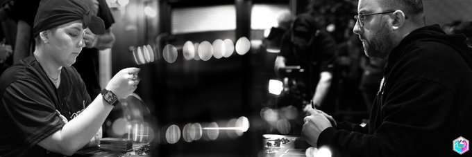 A black and white photo shows two people sitting across from each other at a table, appearing to play a card game in a dimly lit venue.