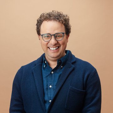 A man with curly hair, glasses, and a wide smile wears a blue blazer over a denim shirt against a peach-colored background. The cheerful, approachable portrait complements the casual, conversational tone of the "same for crypto" comment, lending a friendly human face to the topic.