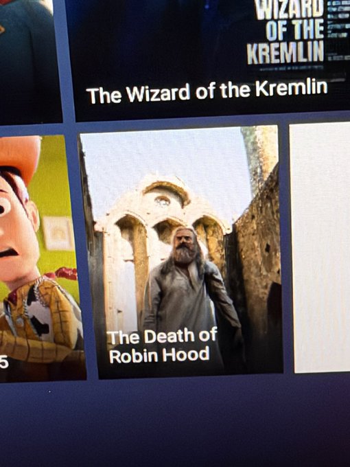 A close-up of an in-flight entertainment screen shows a movie or show titled "The Death of Robin Hood" featuring a bearded man in a gray coat standing in front of stone arches, alongside another option titled "The Wizard of the Kremlin" and a partial image of Woody from Toy Story. The screenshot captures the user's surprise at seeing such a bluntly named film while browsing options during their flight.