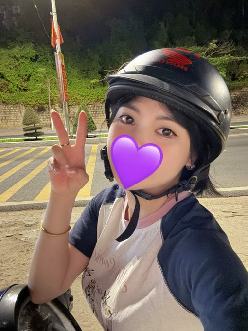 A selfie of a woman wearing a black motorcycle helmet and a beige and navy shirt, making a peace sign with a purple heart emoji covering her mouth.