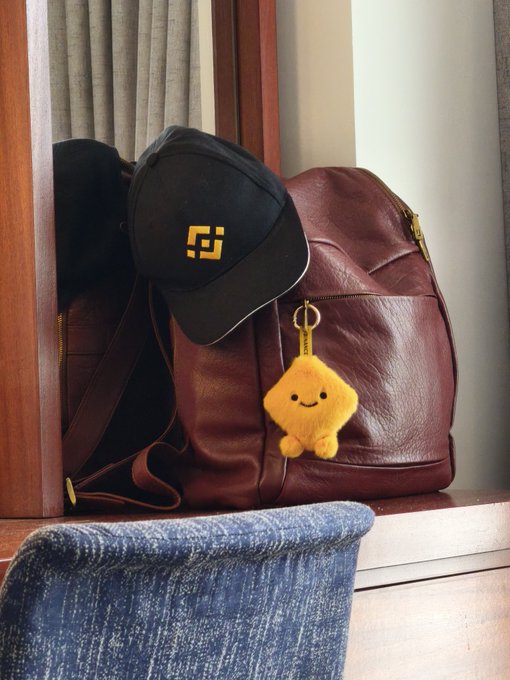 A photograph of a dark maroon leather backpack on a blue textured chair, with a black baseball cap bearing a yellow logo and a small yellow plush star keychain hanging from the zipper.