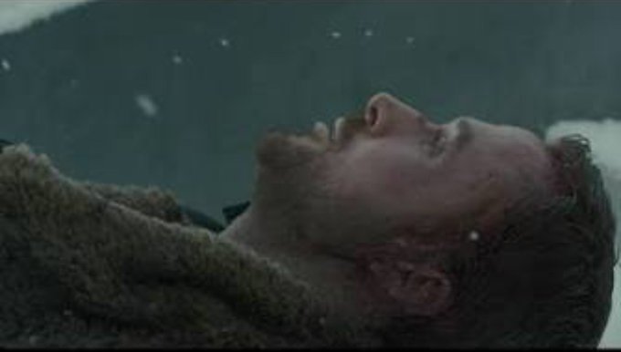 A cinematic close-up of a bearded man lying unconscious in the snow, his face and beard dusted with snowflakes, wearing a heavy knit sweater, suggesting a dramatic or tragic scene.