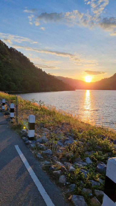A scenic sunset view over a calm lake bordered by forested hills, with a paved roadside in the foreground featuring black-and-white safety posts and rocky embankments.