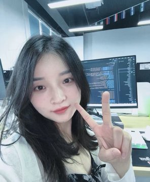 A selfie of a young woman with long dark hair making a peace sign in what appears to be an office with computer monitors visible in the background.