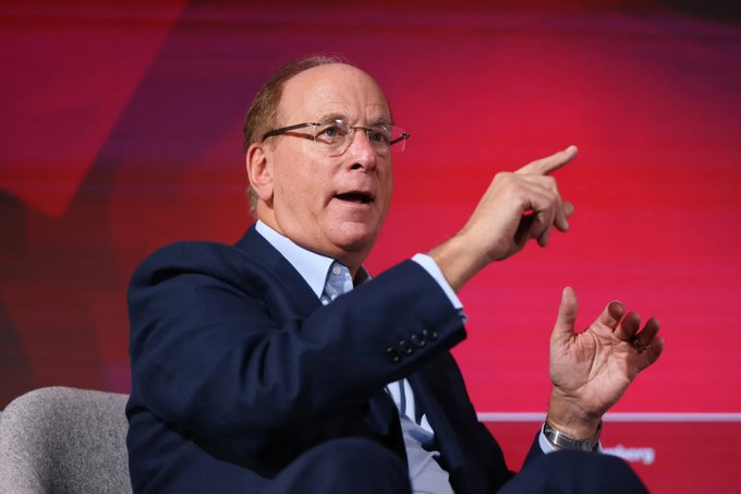 The image features a man dressed in a suit, seated indoors, possibly at an event or conference. He has his hand extended outward, pointing towards something in the distance. In the background behind him, there's a vibrant red wall that serves as a backdrop for the main subjects of the image – the man and the objects he is gesturing with. 

The caption provided also seems to be related to a discussion or presentation taking place at an indoor event. It reads, "BlackRock’s Bitcoin ETF lost $980M in a massive one-week selling streak. This is one of the biggest outflows of the year as big investors pull money out of crypto and into AI stocks." The man's gesture and clothing suggest that he might be discussing a business or financial topic related to cryptocurrency, particularly Bitcoin.