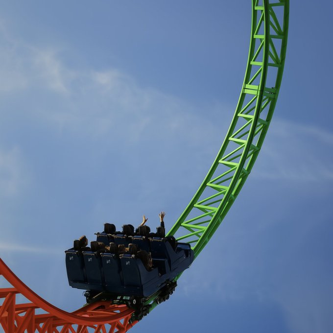A roller coaster car with passengers throwing their hands up as it descends along a bright green track against a blue sky.
