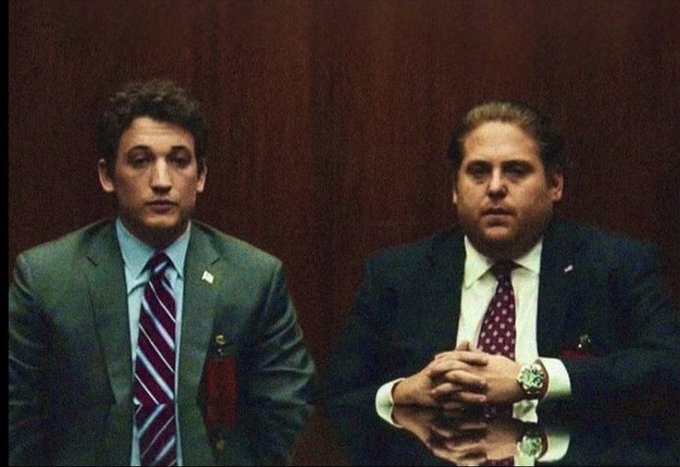 Two men in suits with American flag lapel pins sit at a polished table in a wood-paneled room, with one wearing a watch and clasped hands while the other looks on with a dejected expression, fitting the "terrible financial decisions" theme.