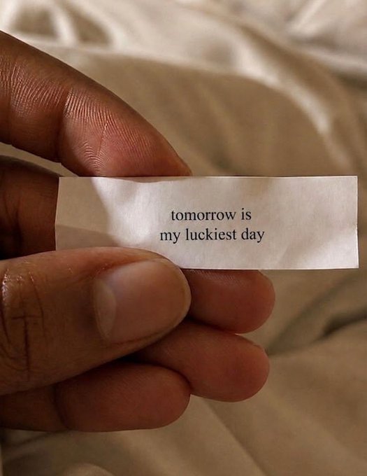 A close-up of a hand holding a small white slip of paper printed with the words 'tomorrow is my luckiest day.'