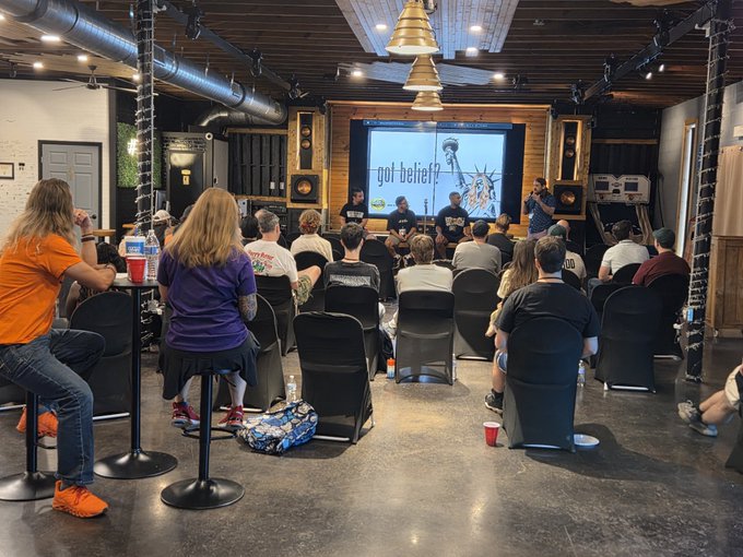 A group of attendees sits facing a presentation screen displaying "got belief?" alongside an image of the Statue of Liberty, at what appears to be the South Carolina SPX6900 conference organized by Country Bitcoin. The event took place in an industrial-style venue with exposed ductwork and pendant lighting, with panelists seated at a table in front of the screen.