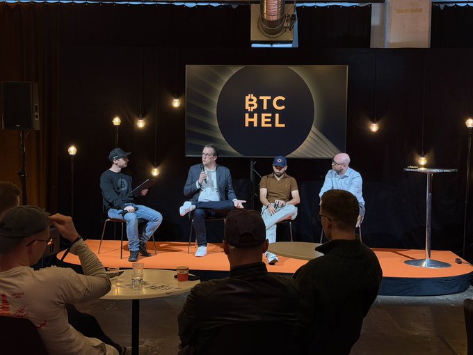 A panel discussion taking place on an orange circular stage, with four seated speakers in front of a large screen displaying the 'BTC HEL' logo, while an audience watches from the foreground.