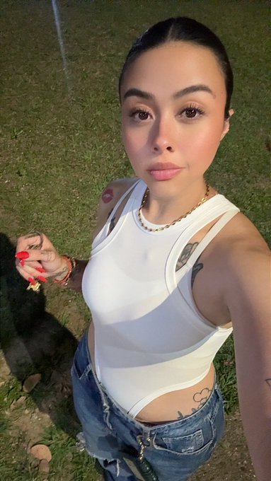 A selfie of a young woman outdoors at night wearing a white tank top and jeans, showing her red manicured nails and visible tattoos.