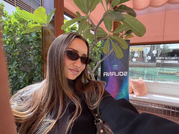 A woman with long brown hair wearing sunglasses takes a selfie in front of a 'SOLANA' banner, with green foliage and a building visible in the background.