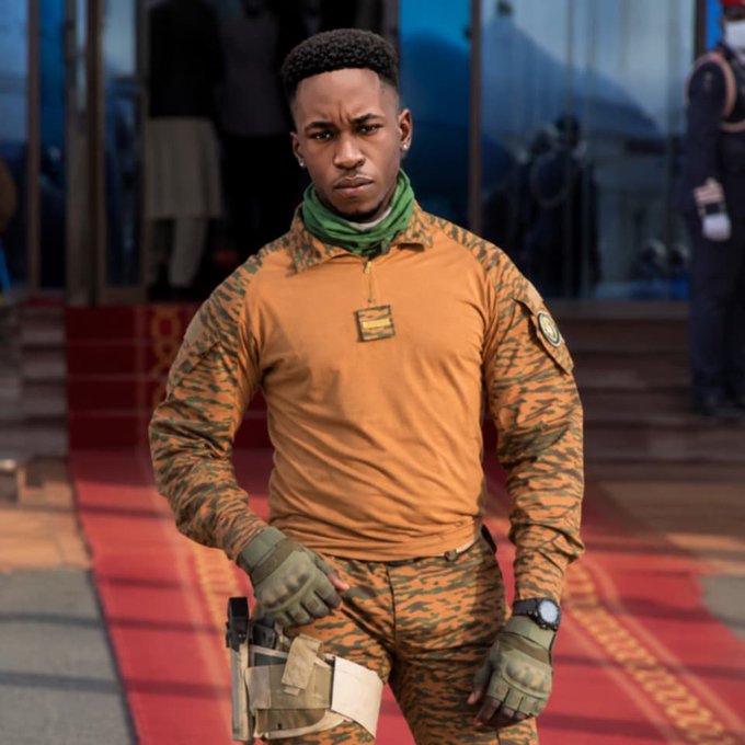A young man with short dark hair wearing an orange and black camouflage-patterned tactical shirt, matching pants, green gloves, and a green neck gaiter, holding a tan holster at his side on a red carpet.