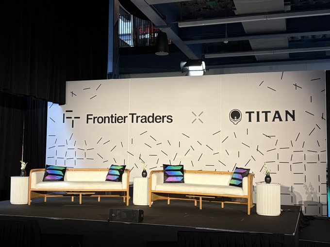 A stage setup with two wooden sofas featuring colorful striped pillows, set against a backdrop displaying 'Frontier Traders' and 'TITAN' logos with a confetti pattern design.