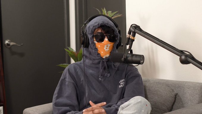 A person wearing a gray hoodie, sunglasses, and an orange bandana over their face sits on a couch with a microphone in front of them, wearing headphones.