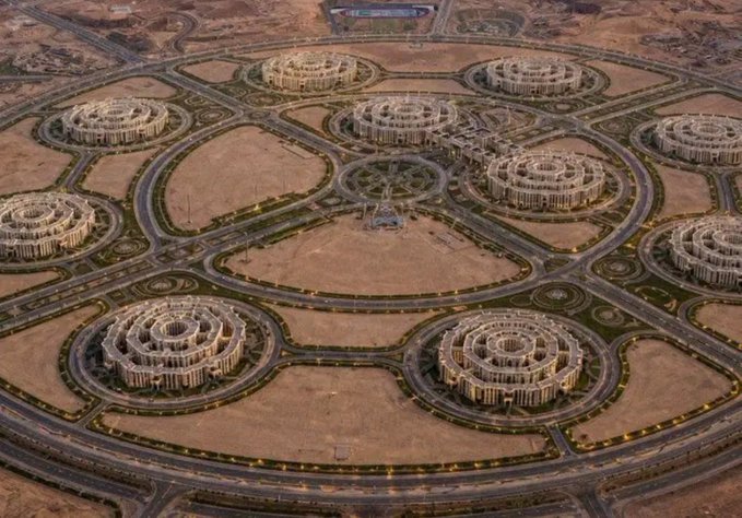 An aerial view of a desert city development featuring multiple identical circular building complexes connected by a network of roads.
