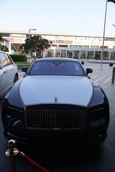 A black and silver Rolls-Royce luxury sedan with the iconic Spirit of Ecstasy hood ornament and chrome grille, parked in front of a modern glass building (appears to be a mall), roped off with a red velvet barrier, with a UAE license plate '76892'.