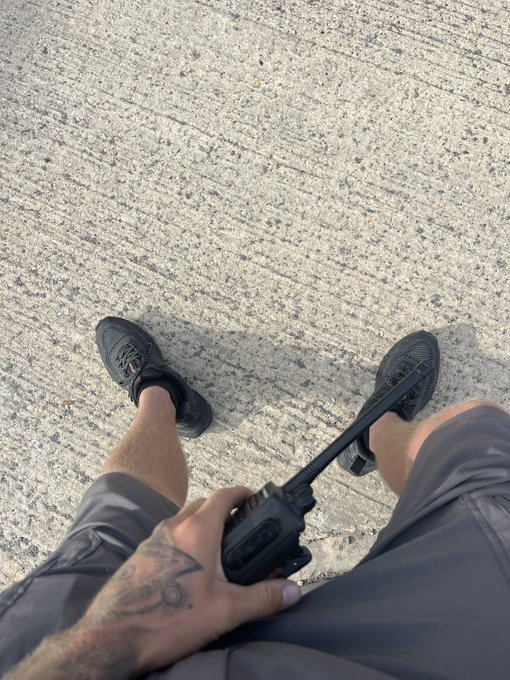 A man with a tattooed hand sits on concrete pavement holding a handheld radio (walkie-talkie), wearing black sneakers and shorts, symbolizing an ordinary working-class individual pursuing the dream of generational financial freedom and retirement for his bloodline ($retire).
