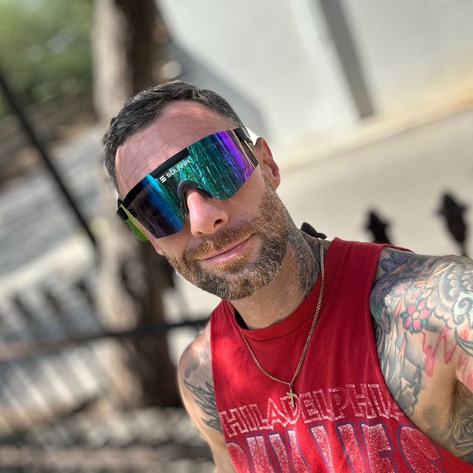 A bearded man with arm tattoos wearing a red tank top and reflective wraparound sunglasses, taking a selfie outdoors near what appears to be a waterfront.