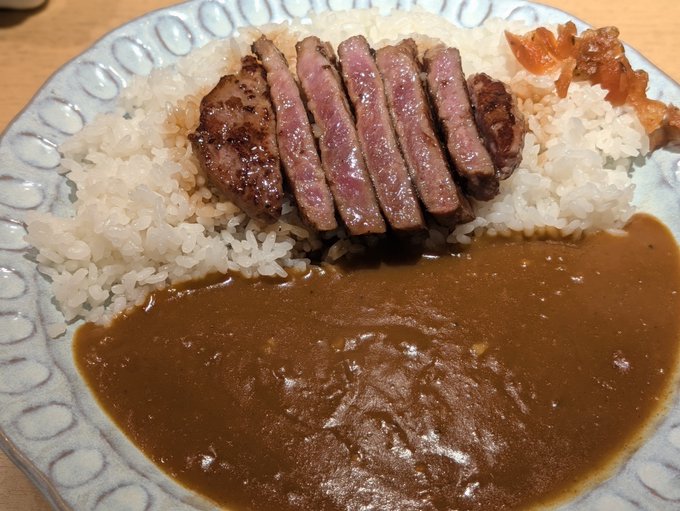 A plate of white rice is topped with sliced medium-rare steak and accompanied by brown curry sauce and a small side of kimchi.