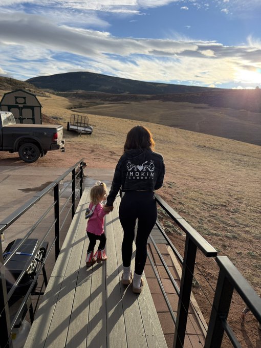 A woman wearing a black 'SMOKIN' jacket holds a small child's hand as they walk down a wooden deck toward a scenic open landscape with rolling hills, a small structure, and a pickup truck under a bright sky.