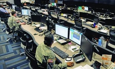 Military personnel in camouflage uniforms working at computer stations in a control room or operations center with multiple monitors.
