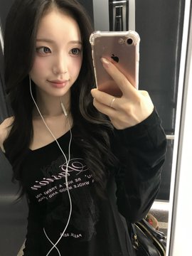 A woman with long dark hair takes a mirror selfie wearing a black top, with white earphones and an Apple iPhone visible in the reflection.