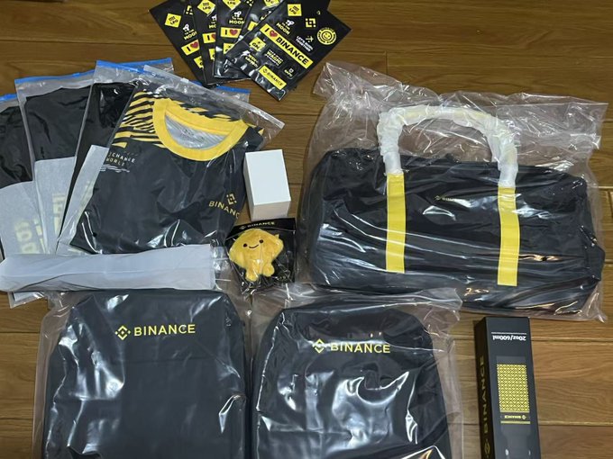 Binance-branded merchandise laid out on a wooden floor, including black and yellow t-shirts, stickers, a small yellow plush figure, and a boxed item.