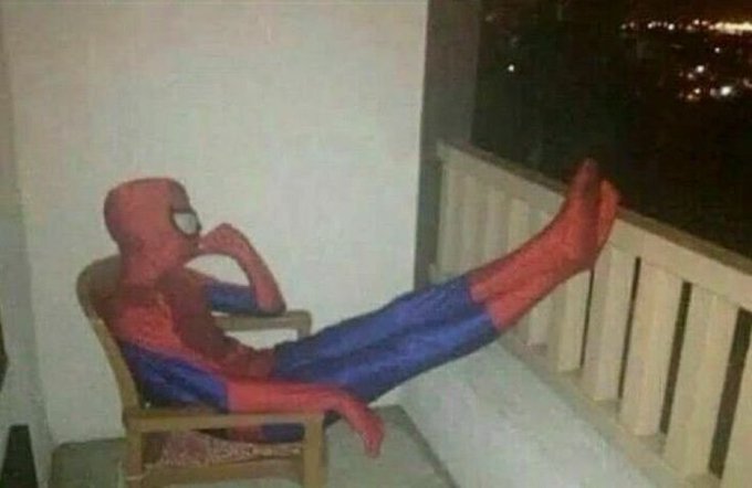 A person dressed in a full Spider-Man costume relaxes in a wooden chair on a balcony at night, with their feet propped up against the railing.