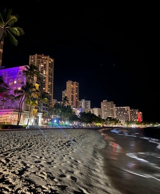 A nighttime photograph of Waikiki Beach in Honolulu, Hawaii, showing the illuminated skyline of high-rise hotels, swaying palm trees, and waves gently lapping the sandy shore under a dark sky.