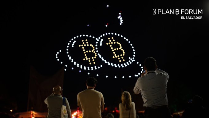 A nighttime drone light show forms the Plan B Forum El Salvador logo above three people watching, with Bitcoin (B) symbols visible in the formation.
