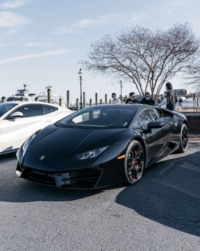 A sleek black Lamborghini Huracán is parked in a lot near a waterfront, with boats visible in the background under a bright blue sky. The image supports the post's claim by visually showcasing the luxury car brand now accepting Bitcoin, symbolizing cryptocurrency's growing influence in high-end markets.