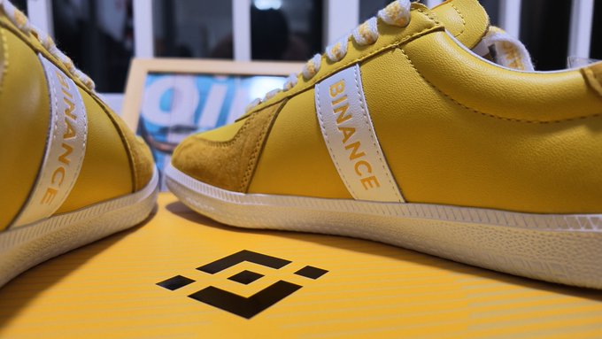 A close-up of yellow sneakers branded with "BINANCE" along the side stripe rests atop a yellow Binance-branded box, playfully tying into the post's joke about "no longer walking barefoot" thanks to Binance-themed footwear.