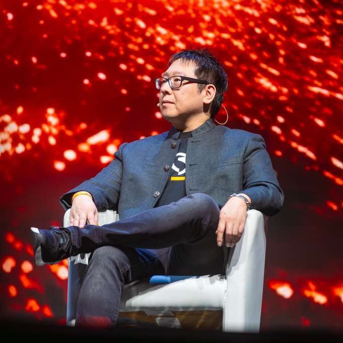 A man with glasses sits cross-legged in a white chair on stage, wearing a dark blazer over a Bitcoin-themed t-shirt, against a dramatic red and orange fiery backdrop. The image aligns with the post's context, showing the featured speaker (@Excellion) at the @LuganoPlanB event, highlighting his return to the Bitcoin-forward city.