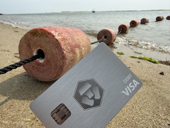 A metal Visa debit card resting on sandy beach near rusted buoy weights and a rope line, with the ocean visible in the background.
