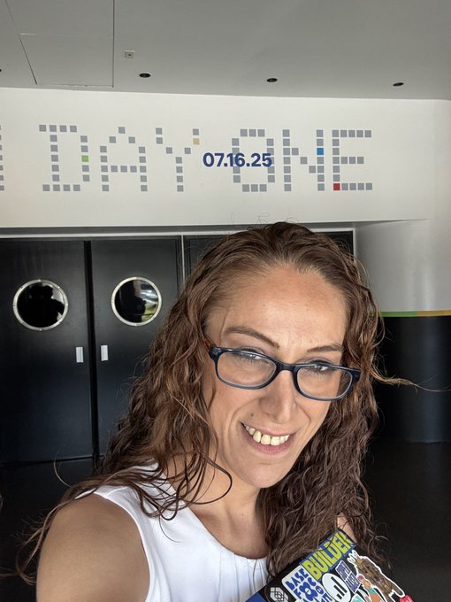 A woman with curly hair and glasses smiles in front of a wall displaying "NXT CODE" and the date "07.16.25," while holding a book titled "The Builder" — tying into the birthday celebration by marking the special date and suggesting a shared appreciation for innovation and community building.