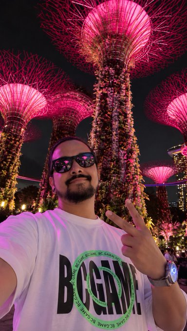 A man wearing a "BECAME" graphic t-shirt and sunglasses throws a peace sign for a selfie at Singapore's iconic Supertree Grove, illuminated in vibrant pink at night—capturing the memorable vibes of Token2049.