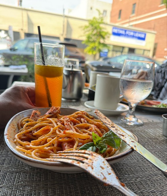 A plate of pasta in tomato sauce garnished with basil, served at an outdoor restaurant table alongside a glass of iced tea, a glass of water, and a coffee cup.