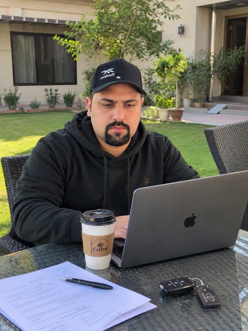 A man wearing a black cap with 'Rajasthan Royals' text and a black hoodie working on an Apple laptop at an outdoor glass table, with a 'The Coffee' cup, keys, and papers nearby.