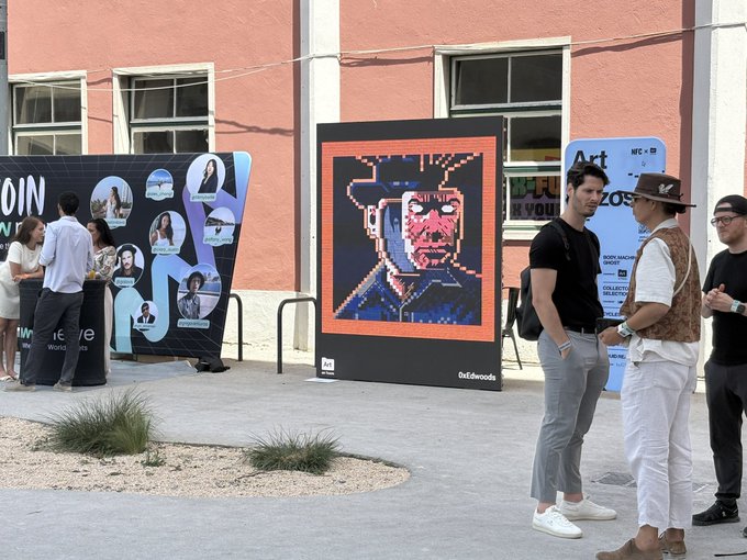 The image features a group of people standing around an art exhibit, with one person wearing a cowboy hat. The main attraction of the exhibit is a large screen displaying a digital artwork featuring a man's face. The backdrop includes a pink building and a sign that reads "@NFC Summit Lisbon".