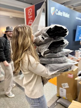 A woman wearing a white sweater is holding a large stack of clothing bags, including jackets and pants, which are neatly packed together. In the background, there's another person who appears to be looking at the pile of clothes. The image also contains text that reads "Official @cakewallet jacket stacker".