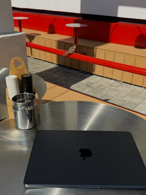 A black Apple MacBook laptop sitting closed on a round metal café table, with a small bird perched on a red railing in the sunny outdoor background.