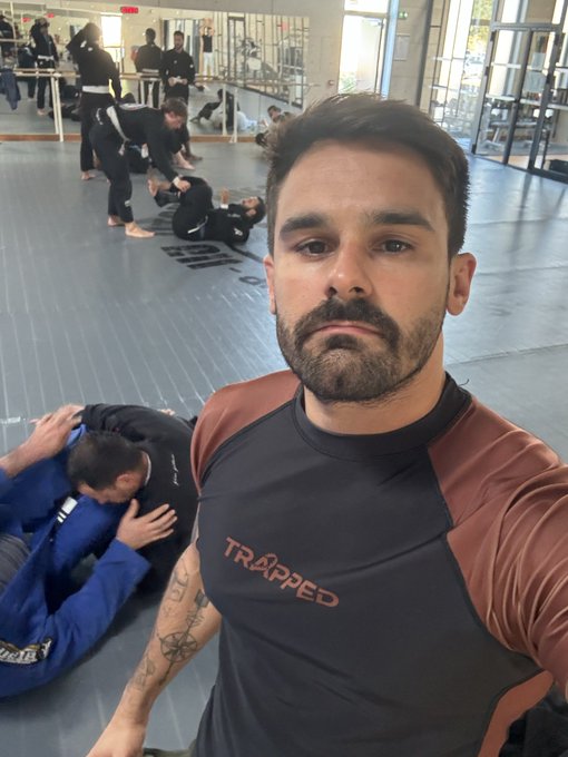 Image 9: A selfie taken inside a martial arts gym showing a man with a beard wearing a rash guard with "TRAPPED" printed on it, while other people practice grappling on the mats in the background.