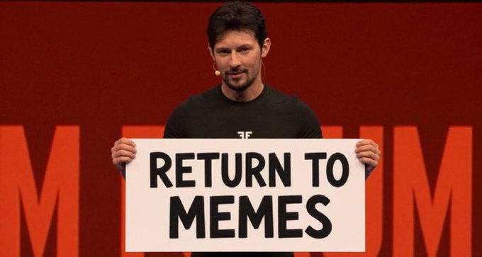 A man stands on a TEDx stage holding a large sign that reads 'RETURN TO MEMES'.