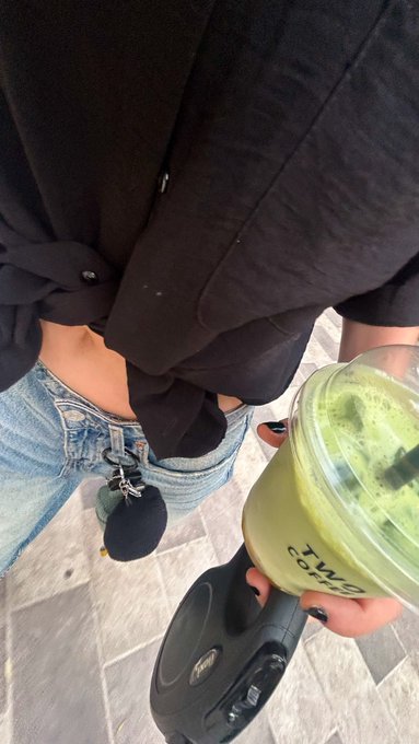 A person looks down at their outfit, showing a black hoodie, blue jeans with keys and a pouch attached, black sneakers, and a green drink in a 'MATCHA' branded cup, while holding a camera rig.