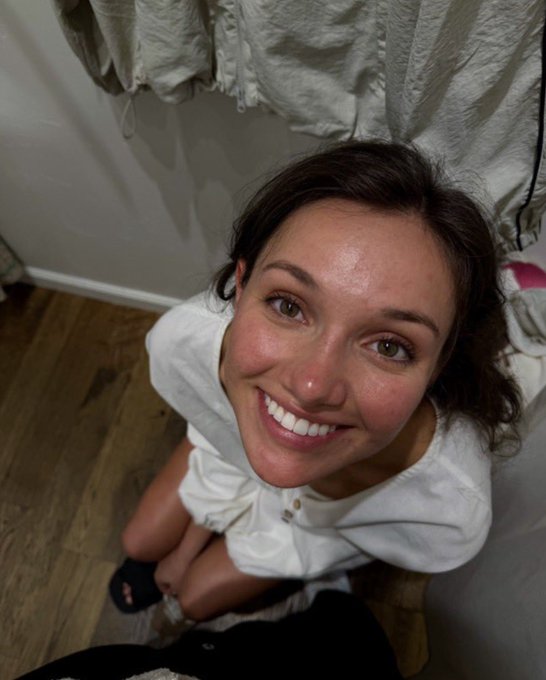A woman with brown hair smiles brightly up at the camera from a high angle, wearing a white top in a room with wooden floors and what appears to be a black cat partially visible.