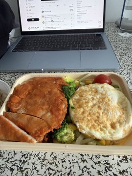 The image shows a meal consisting of a chicken dish, an egg dish, and vegetables such as broccoli and carrots. The food items are placed inside a container or tray, which is sitting on top of a table. A laptop computer is also present in the scene, suggesting that someone might be working while enjoying their meal.
