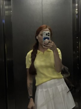 A mirror selfie taken inside an elevator showing a woman with red braided hair wearing a yellow polo shirt and white skirt.
