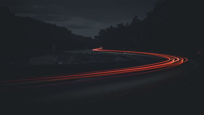 A long-exposure night photograph of a winding road featuring bright red tail light trails curving through a dark landscape.