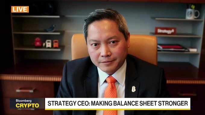 The image features a man wearing an orange tie, sitting behind a desk with his hands on his lap. The text "BOOMBERRY" can be seen at the top of the image, which may suggest that he is a leader or expert in the field. The man appears to be talking about strategy and balance sheets, possibly discussing how these concepts apply to Bitcoin leadership.